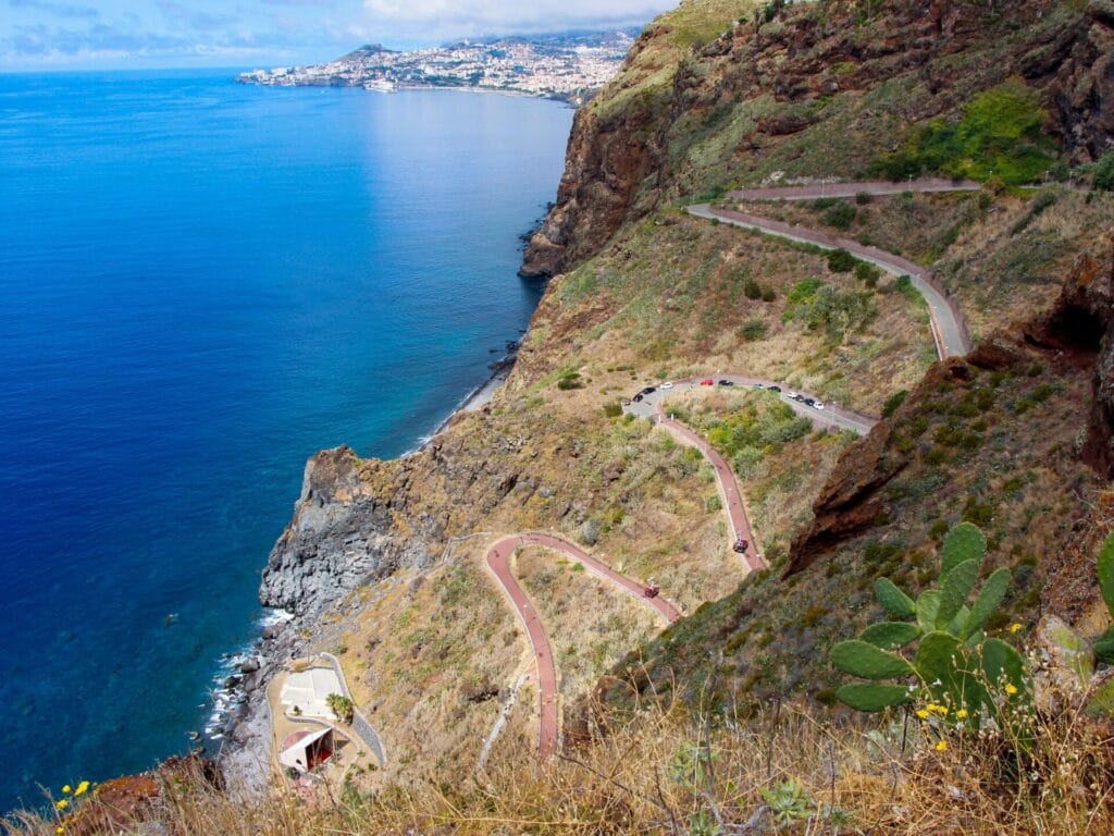 Eine kurvenreiche Strasse windet sich an der Steilküste zum Meer hinunter. Im Hintergrund ist eine Stadt zu sehen.