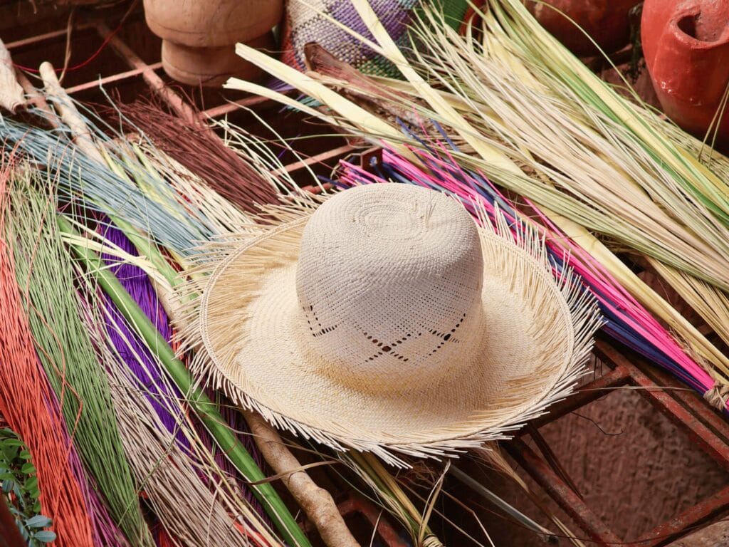 Ein naturfarbener Strohhut, umrahmt von gefärbten und naturfarbenen Palmblättern. Im Hintergrund sind Teile von Tontöpfen zu sehen.